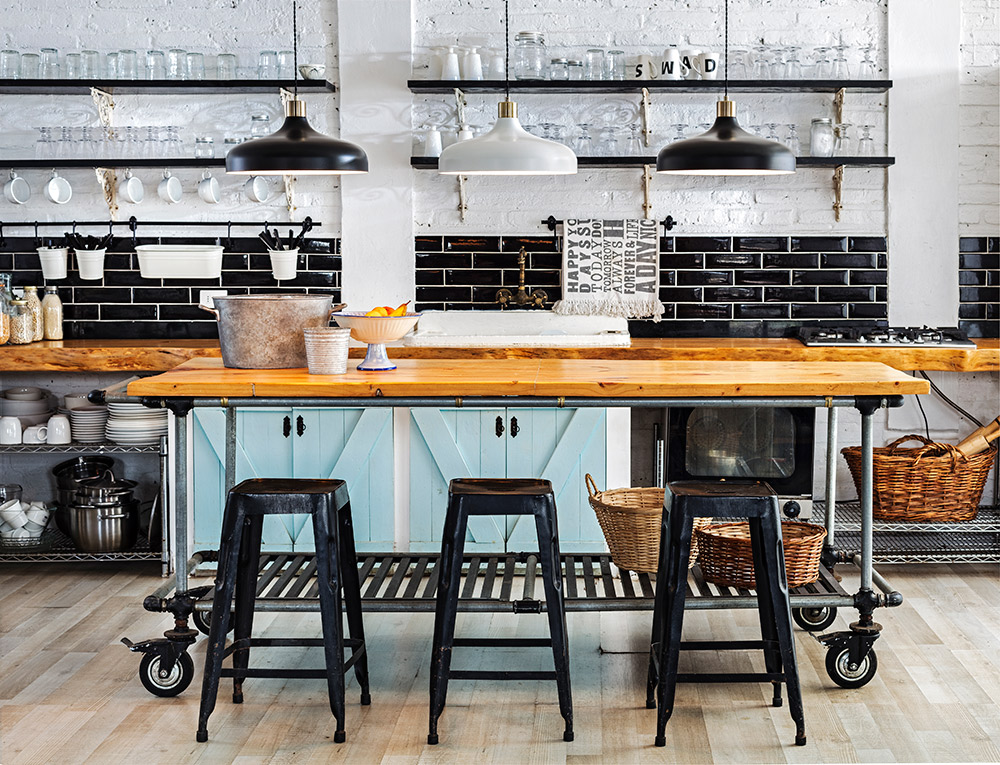 Imagen de comedor con decoración industrial, mesa con ruedas y sillas metálicas color negro tipo bar. Se ven 3 lámparas colgantes como iluminación